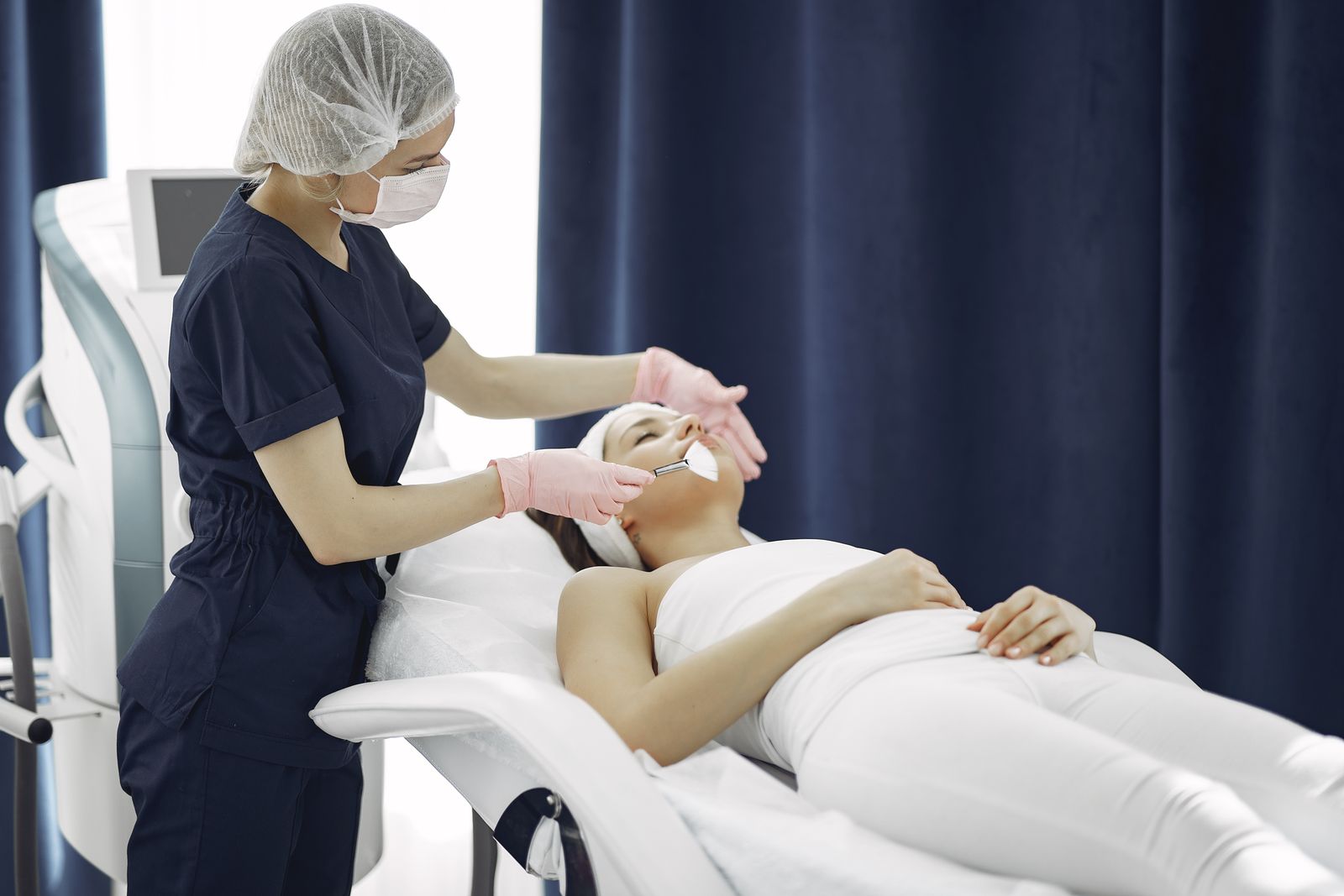 Aesthetician applying a facial mask to a woman in a treatment room.